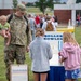 A military service member in camouflage uniform holds a young infant while standing near a 'Roller Bowler' carnival game at an outdoor family event. Two girls with their backs to the camera appear to be playing or observing the game. The setting appears to be a military base family day or community event on a grassy field.
