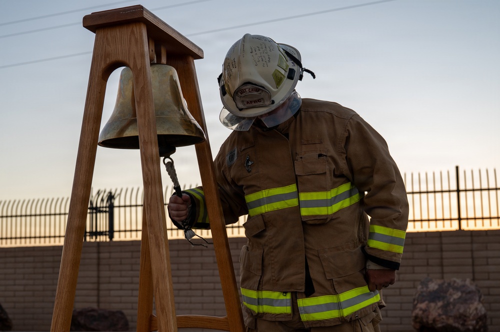 A firefighter in full turnout gear is ringing a brass memorial bell mounted on a wooden stand. The helmet is marked with 'BAT CHIEF' and 'AFRC' insignia, suggesting this is a battalion chief from an Air Force Reserve Command fire department. The setting appears to be an outdoor ceremony, likely a memorial or remembrance event, photographed at dusk or dawn.
