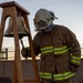 A firefighter in full turnout gear is ringing a brass memorial bell mounted on a wooden stand. The helmet is marked with 'BAT CHIEF' and 'AFRC' insignia, suggesting this is a battalion chief from an Air Force Reserve Command fire department. The setting appears to be an outdoor ceremony, likely a memorial or remembrance event, photographed at dusk or dawn.