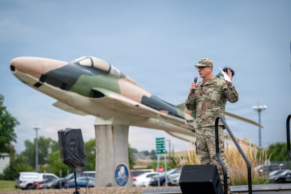 A military officer in OCP (Operational Camouflage Pattern) uniform stands on a stage speaking into a microphone at an outdoor ceremony. Behind him is a static display aircraft mounted on a pedestal, painted in a camouflage scheme of tan and green, consistent with Vietnam-era or Cold War-era jet aircraft. The setting appears to be a military installation entrance or memorial area.