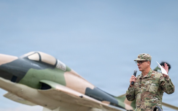 A military officer in OCP (Operational Camouflage Pattern) uniform stands on a stage speaking into a microphone at an outdoor ceremony. Behind him is a static display aircraft mounted on a pedestal, painted in a camouflage scheme of tan and green, consistent with Vietnam-era or Cold War-era jet aircraft. The setting appears to be a military installation entrance or memorial area.