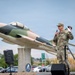 A military officer in OCP (Operational Camouflage Pattern) uniform stands on a stage speaking into a microphone at an outdoor ceremony. Behind him is a static display aircraft mounted on a pedestal, painted in a camouflage scheme of tan and green, consistent with Vietnam-era or Cold War-era jet aircraft. The setting appears to be a military installation entrance or memorial area.