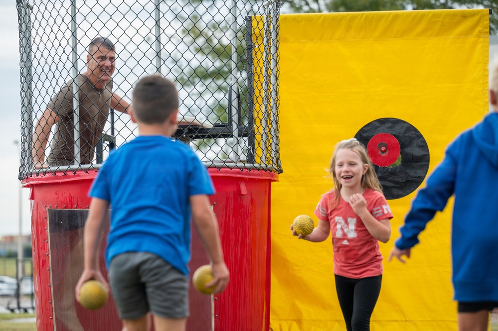 Children are participating in a dunk tank activity at what appears to be an outdoor fair or community event. A man sits in the elevated cage of a red dunk tank while kids hold throwing balls and attempt to hit the circular target mounted on the yellow backdrop. A girl in a red Nebraska 'N' shirt is laughing and appears to have just thrown a ball.