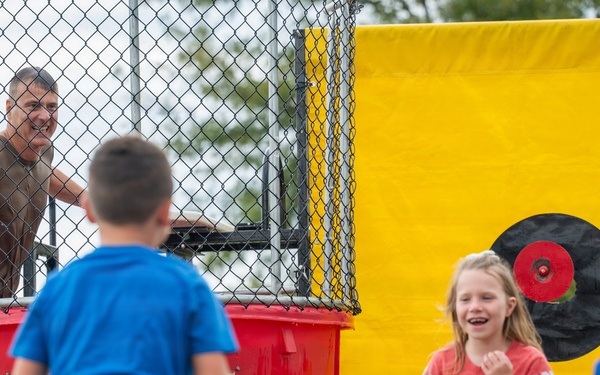Children are participating in a dunk tank activity at what appears to be an outdoor fair or community event. A man sits in the elevated cage of a red dunk tank while kids hold throwing balls and attempt to hit the circular target mounted on the yellow backdrop. A girl in a red Nebraska 'N' shirt is laughing and appears to have just thrown a ball.