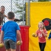 Children are participating in a dunk tank activity at what appears to be an outdoor fair or community event. A man sits in the elevated cage of a red dunk tank while kids hold throwing balls and attempt to hit the circular target mounted on the yellow backdrop. A girl in a red Nebraska 'N' shirt is laughing and appears to have just thrown a ball.
