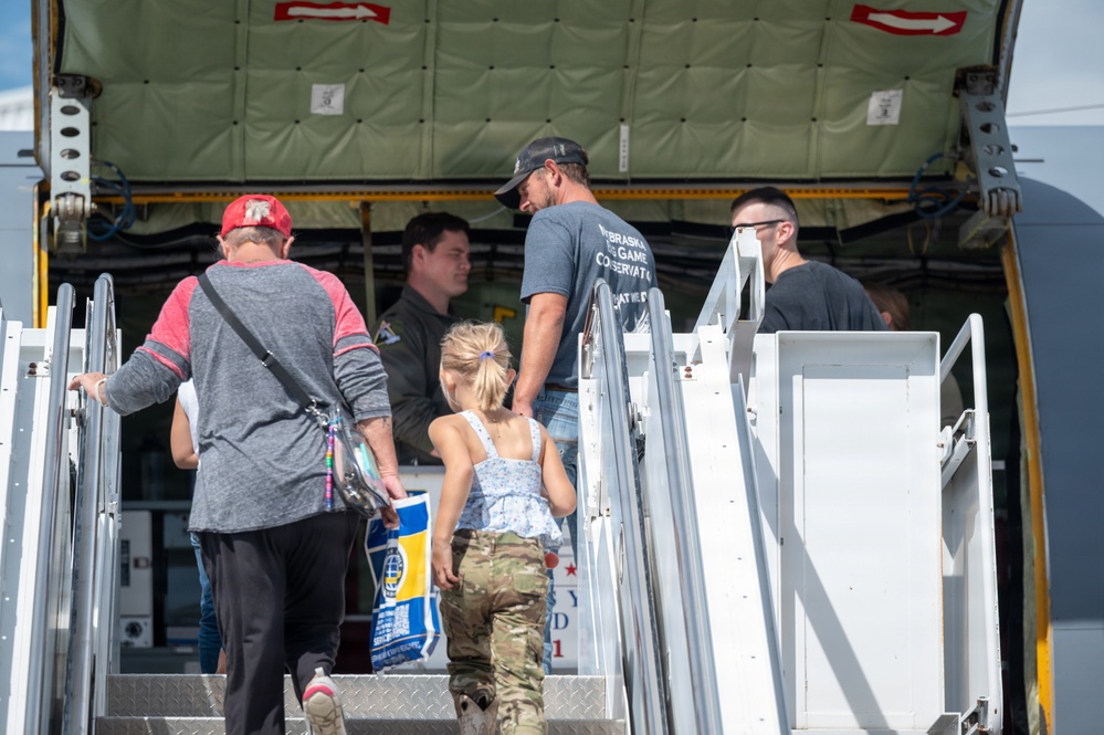 A group of people, including adults and a young child, are boarding or touring a large military aircraft via a mobile staircase. A uniformed crew member is visible at the aircraft entrance greeting or guiding visitors. The setting appears to be an airshow or open house event based on the civilian attire and informal atmosphere.