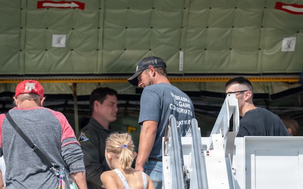 A group of people, including adults and a young child, are boarding or touring a large military aircraft via a mobile staircase. A uniformed crew member is visible at the aircraft entrance greeting or guiding visitors. The setting appears to be an airshow or open house event based on the civilian attire and informal atmosphere.