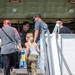 A group of people, including adults and a young child, are boarding or touring a large military aircraft via a mobile staircase. A uniformed crew member is visible at the aircraft entrance greeting or guiding visitors. The setting appears to be an airshow or open house event based on the civilian attire and informal atmosphere.