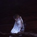 A barn owl (Tyto alba) is perched on a wooden branch in a very dark environment, illuminated by a focused light source that highlights its distinctive white and speckled plumage. The owl appears to be in a resting or preening posture with its head bowed downward and one wing partially extended below the branch. The dark wooden background suggests an indoor enclosure or barn-like structure.