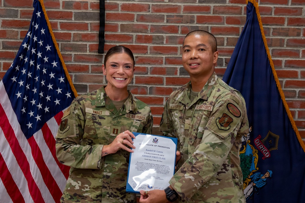 Two U.S. Air Force personnel are posing together holding a Certificate of Promotion to the rank of Airman First Class for 'Randell M. Luterte.' The ceremony appears to take place in front of an American flag and what appears to be the Maine state flag (featuring the word 'Dirigo'). Both individuals are wearing OCP (Operational Camouflage Pattern) uniforms.
