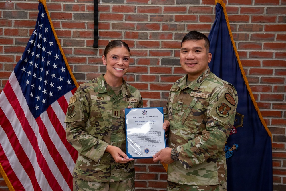 Two U.S. Air Force personnel in OCP (Operational Camouflage Pattern) uniforms are posing together while holding a Certificate of Promotion in front of an American flag and a blue military unit flag against a brick wall backdrop. The female service member on the left, whose name tape reads 'AWALT,' appears to hold the rank of Captain (visible rank insignia). This appears to be an official military promotion ceremony photo.