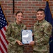 Two U.S. Air Force personnel in OCP (Operational Camouflage Pattern) uniforms are posing together while holding a Certificate of Promotion in front of an American flag and a blue military unit flag against a brick wall backdrop. The female service member on the left, whose name tape reads 'AWALT,' appears to hold the rank of Captain (visible rank insignia). This appears to be an official military promotion ceremony photo.