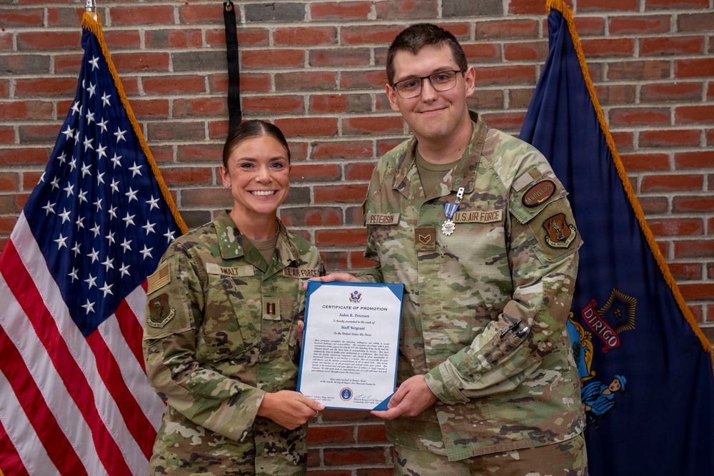 Two U.S. Air Force personnel pose together holding a Certificate of Promotion. The male airman, identified by his name tape as 'Peterson,' has been promoted to Staff Sergeant and wears a medal on his uniform. They are standing in front of an American flag and what appears to be a Maine state flag against a brick wall backdrop.