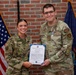 Two U.S. Air Force personnel pose together holding a Certificate of Promotion. The male airman, identified by his name tape as 'Peterson,' has been promoted to Staff Sergeant and wears a medal on his uniform. They are standing in front of an American flag and what appears to be a Maine state flag against a brick wall backdrop.
