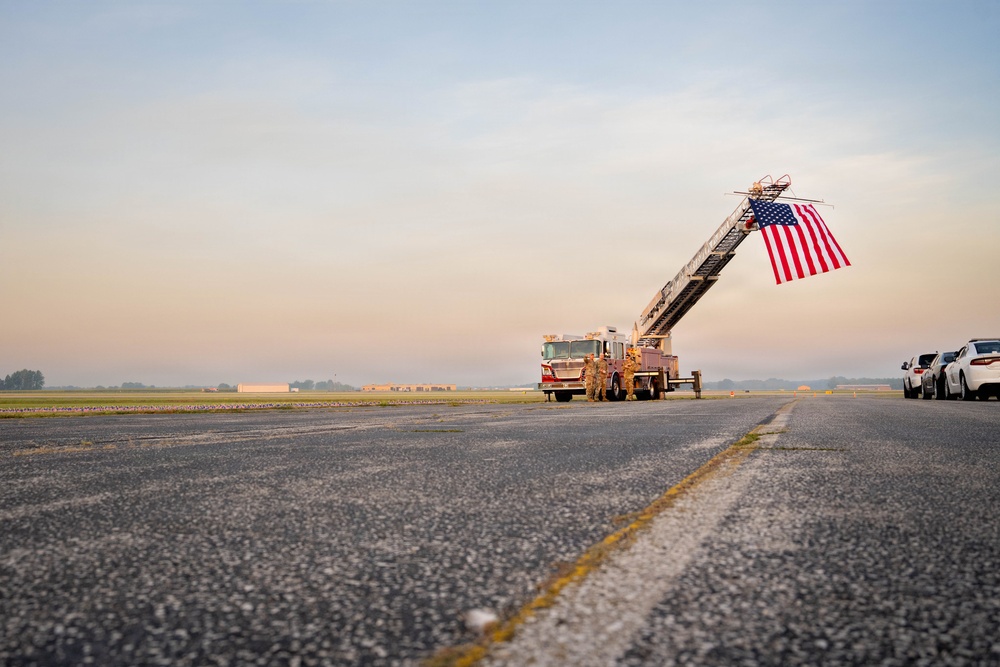 A fire truck with an extended aerial ladder displaying a large American flag stands on what appears to be an airport tarmac or airfield at dawn or dusk. Several individuals in uniform are standing near the truck, and a line of civilian vehicles is parked to the right. The scene suggests a ceremonial or memorial event, likely a dignified transfer or honor guard ceremony.
