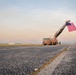 A fire truck with an extended aerial ladder displaying a large American flag stands on what appears to be an airport tarmac or airfield at dawn or dusk. Several individuals in uniform are standing near the truck, and a line of civilian vehicles is parked to the right. The scene suggests a ceremonial or memorial event, likely a dignified transfer or honor guard ceremony.
