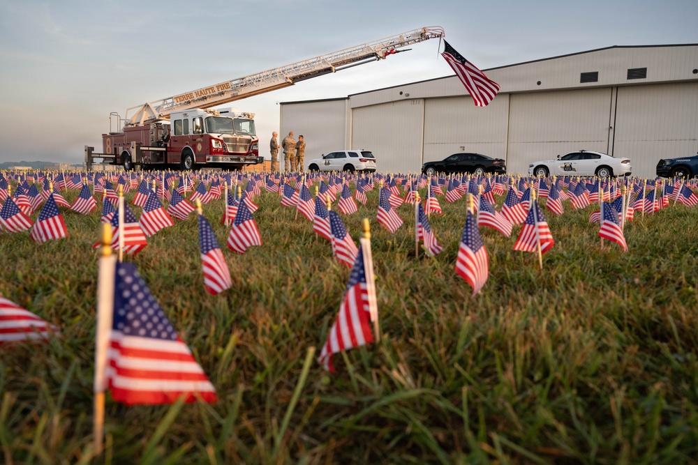 A large field of small American flags planted in grass serves as a memorial display, likely commemorating a patriotic or remembrance event. A Terre Haute Fire Department ladder truck is visible in the background with its aerial ladder extended and an American flag flying from the top. A large hangar building, several vehicles, and a few individuals in military-style uniforms are also present in the background.