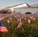 A large field of small American flags planted in grass serves as a memorial display, likely commemorating a patriotic or remembrance event. A Terre Haute Fire Department ladder truck is visible in the background with its aerial ladder extended and an American flag flying from the top. A large hangar building, several vehicles, and a few individuals in military-style uniforms are also present in the background.