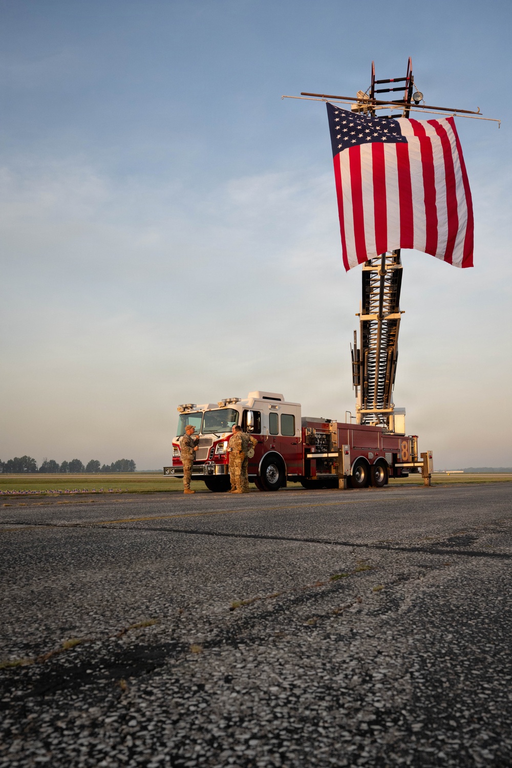 A red ladder fire truck with the number '8' visible is parked on what appears to be a military airfield tarmac, with its aerial ladder extended and displaying a large American flag. Two individuals in military camouflage uniforms stand near the front of the truck. The scene is photographed during golden hour lighting with a flat, open landscape visible in the background.