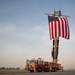 A red ladder fire truck with the number '8' visible is parked on what appears to be a military airfield tarmac, with its aerial ladder extended and displaying a large American flag. Two individuals in military camouflage uniforms stand near the front of the truck. The scene is photographed during golden hour lighting with a flat, open landscape visible in the background.