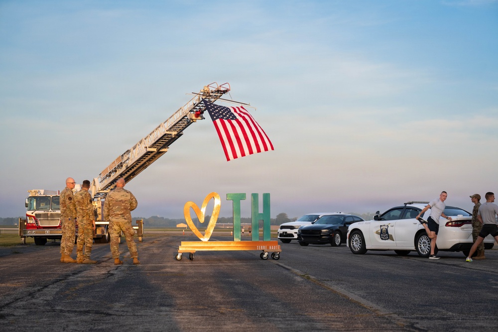 A welcome or homecoming ceremony is taking place on an airport tarmac in Terre Haute, Indiana. A fire truck ladder is extended at an angle displaying a large American flag, while military personnel in camouflage uniforms stand nearby a decorative 'TH' (Terre Haute) sign. Law enforcement vehicles and civilians are also present on the tarmac.