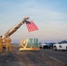 A welcome or homecoming ceremony is taking place on an airport tarmac in Terre Haute, Indiana. A fire truck ladder is extended at an angle displaying a large American flag, while military personnel in camouflage uniforms stand nearby a decorative 'TH' (Terre Haute) sign. Law enforcement vehicles and civilians are also present on the tarmac.