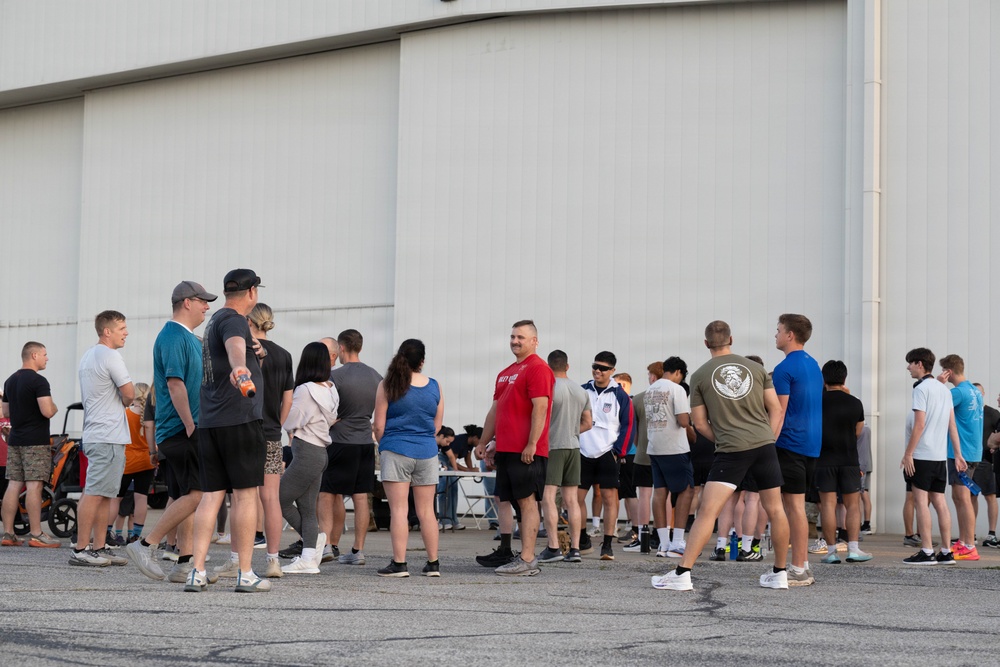 A large group of people, dressed in casual athletic wear, are gathered outside in front of a large industrial or hangar-style building. They appear to be assembled for an organized outdoor event, possibly a fun run, fitness event, or community gathering. A registration or check-in table is visible in the background.
