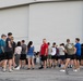 A large group of people, dressed in casual athletic wear, are gathered outside in front of a large industrial or hangar-style building. They appear to be assembled for an organized outdoor event, possibly a fun run, fitness event, or community gathering. A registration or check-in table is visible in the background.