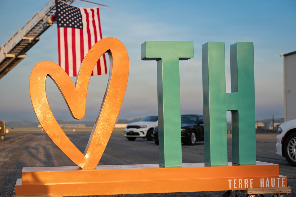 A decorative civic sign featuring a large orange/gold heart symbol alongside green block letters 'TH' (representing Terre Haute) mounted on an orange base platform. The sign is positioned outdoors on what appears to be a tarmac or airfield, with an American flag displayed from a fire truck ladder in the background. The watermark 'TERRE HAUTE' is visible in the lower right corner, confirming this is a promotional or civic installation for Terre Haute, Indiana.