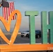 A decorative civic sign featuring a large orange/gold heart symbol alongside green block letters 'TH' (representing Terre Haute) mounted on an orange base platform. The sign is positioned outdoors on what appears to be a tarmac or airfield, with an American flag displayed from a fire truck ladder in the background. The watermark 'TERRE HAUTE' is visible in the lower right corner, confirming this is a promotional or civic installation for Terre Haute, Indiana.