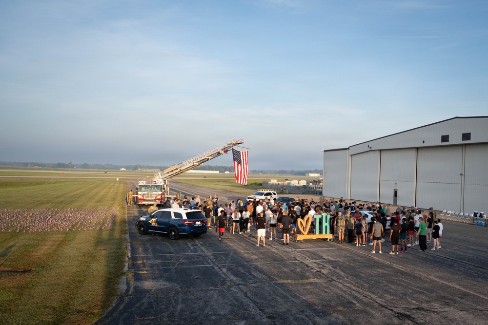 A patriotic gathering at a small regional airport or airfield, where a large crowd of people has assembled near a hangar. A fire truck has extended its aerial ladder to display a large American flag hanging vertically. The scene appears to be a community welcome or memorial event, suggested by the decorative signage and rows of small flags planted in the grass.