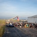 A patriotic gathering at a small regional airport or airfield, where a large crowd of people has assembled near a hangar. A fire truck has extended its aerial ladder to display a large American flag hanging vertically. The scene appears to be a community welcome or memorial event, suggested by the decorative signage and rows of small flags planted in the grass.
