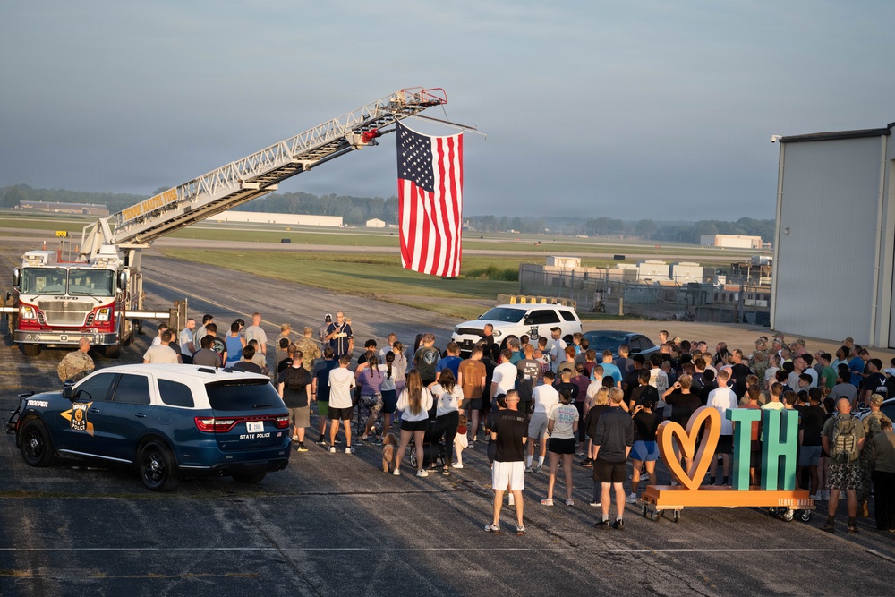A patriotic outdoor gathering is taking place on what appears to be a military airfield or airport tarmac. A large American flag is suspended from an extended fire truck ladder, while a crowd of approximately 100+ people assembles below, accompanied by law enforcement and emergency vehicles. A decorative sign reading 'TH' with a heart symbol is visible on the right side, suggesting a community or civic event.
