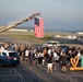 A patriotic outdoor gathering is taking place on what appears to be a military airfield or airport tarmac. A large American flag is suspended from an extended fire truck ladder, while a crowd of approximately 100+ people assembles below, accompanied by law enforcement and emergency vehicles. A decorative sign reading 'TH' with a heart symbol is visible on the right side, suggesting a community or civic event.