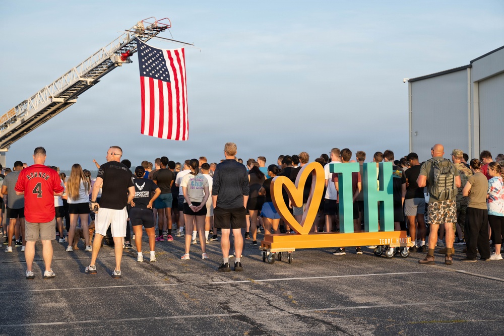 A large outdoor gathering is taking place on what appears to be a tarmac or airfield, with a crowd of people assembled near a decorative 'Love TH' (Terre Haute) sign. An American flag is prominently displayed hanging from a fire truck ladder aerial boom overhead. The event appears to be a community or military-affiliated gathering, possibly a ceremony or commemorative run/event.