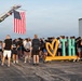 A large outdoor gathering is taking place on what appears to be a tarmac or airfield, with a crowd of people assembled near a decorative 'Love TH' (Terre Haute) sign. An American flag is prominently displayed hanging from a fire truck ladder aerial boom overhead. The event appears to be a community or military-affiliated gathering, possibly a ceremony or commemorative run/event.