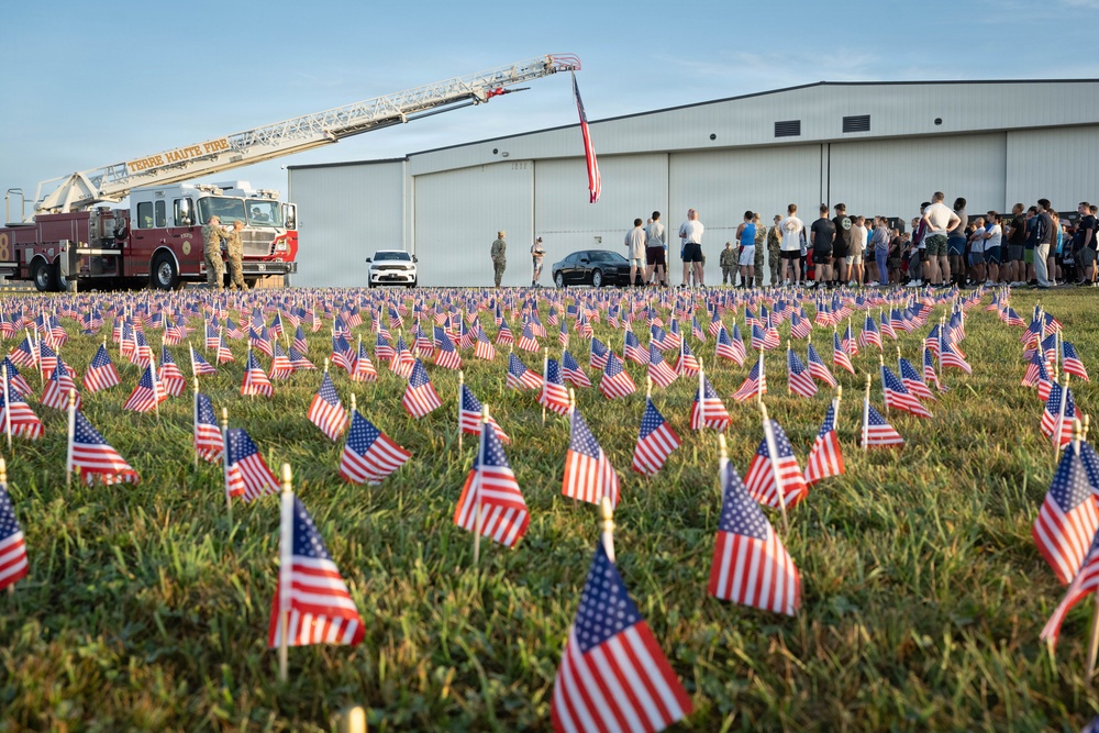 A memorial scene featuring hundreds of small American flags planted in a grass field, likely commemorating a patriotic event such as 9/11 or Memorial Day. A Terre Haute Fire Department ladder truck is visible with its aerial ladder extended and an American flag hanging from it. A group of military personnel and civilians are gathered near a large hangar building in the background.