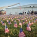 A memorial scene featuring hundreds of small American flags planted in a grass field, likely commemorating a patriotic event such as 9/11 or Memorial Day. A Terre Haute Fire Department ladder truck is visible with its aerial ladder extended and an American flag hanging from it. A group of military personnel and civilians are gathered near a large hangar building in the background.