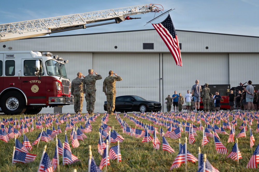 A solemn military memorial ceremony is taking place near an aircraft hangar, with soldiers in camouflage uniforms saluting a large American flag suspended from a fire truck's extended aerial ladder. The foreground is filled with hundreds of small American flags planted in the grass, suggesting a tribute to fallen service members. A fire truck marked 'THFD' and a group of onlookers are visible in the background.