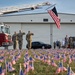 A solemn military memorial ceremony is taking place near an aircraft hangar, with soldiers in camouflage uniforms saluting a large American flag suspended from a fire truck's extended aerial ladder. The foreground is filled with hundreds of small American flags planted in the grass, suggesting a tribute to fallen service members. A fire truck marked 'THFD' and a group of onlookers are visible in the background.