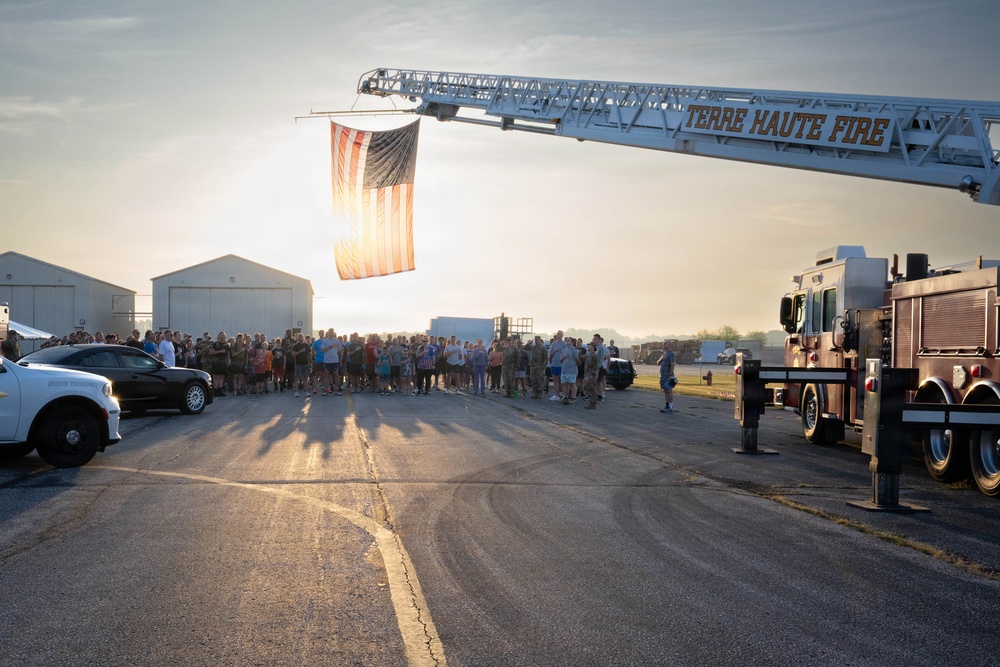 A large group of people, including what appear to be military personnel and civilians, are gathered on a tarmac or airfield in Terre Haute, Indiana. A Terre Haute Fire Department ladder truck has extended its aerial ladder to display a large American flag backlit by a low sun. The scene suggests a ceremonial or commemorative event, possibly a memorial run, sendoff, or patriotic gathering.