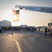 A large group of people, including what appear to be military personnel and civilians, are gathered on a tarmac or airfield in Terre Haute, Indiana. A Terre Haute Fire Department ladder truck has extended its aerial ladder to display a large American flag backlit by a low sun. The scene suggests a ceremonial or commemorative event, possibly a memorial run, sendoff, or patriotic gathering.