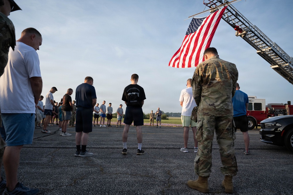 A group of military personnel and civilians are gathered outdoors on a paved surface in what appears to be a memorial or solemn ceremony. An American flag is displayed prominently from a fire truck ladder in the upper right of the frame. Participants have their heads bowed, suggesting a moment of silence or prayer.
