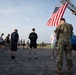 A group of military personnel and civilians are gathered outdoors on a paved surface in what appears to be a memorial or solemn ceremony. An American flag is displayed prominently from a fire truck ladder in the upper right of the frame. Participants have their heads bowed, suggesting a moment of silence or prayer.