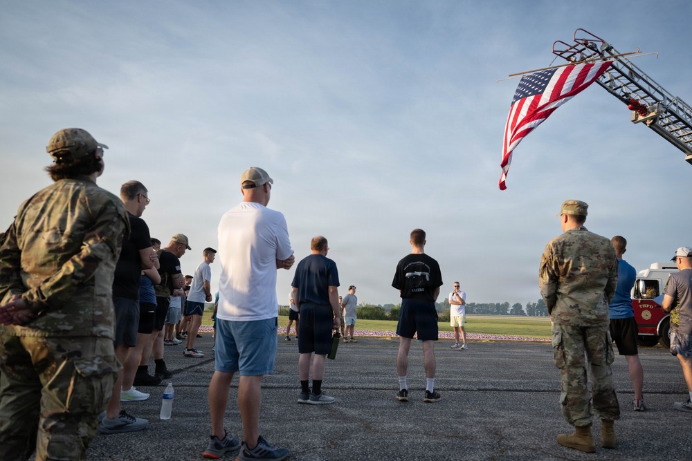 A group of military personnel and civilians are gathered on a tarmac or airfield in what appears to be a commemorative or memorial event. An American flag is displayed from a fire department ladder truck (marked 'THFD') in the upper right corner. Small flags or markers are visible in the background field, suggesting a patriotic or remembrance ceremony.