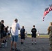 A group of military personnel and civilians are gathered on a tarmac or airfield in what appears to be a commemorative or memorial event. An American flag is displayed from a fire department ladder truck (marked 'THFD') in the upper right corner. Small flags or markers are visible in the background field, suggesting a patriotic or remembrance ceremony.