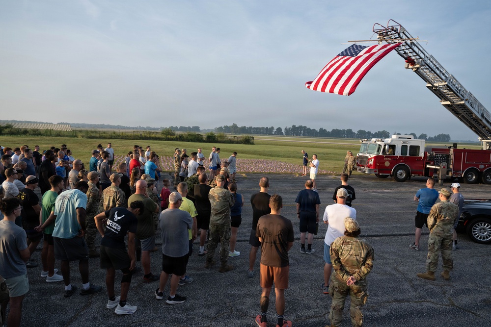 A large group of military personnel and civilians are gathered outdoors on a tarmac or airfield for what appears to be a memorial or commemorative event. A large American flag is displayed from an extended fire truck ladder on the right side of the image. In the background, a field of small flags is visible, suggesting a tribute or remembrance ceremony.