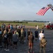 A large group of military personnel and civilians are gathered outdoors on a tarmac or airfield for what appears to be a memorial or commemorative event. A large American flag is displayed from an extended fire truck ladder on the right side of the image. In the background, a field of small flags is visible, suggesting a tribute or remembrance ceremony.