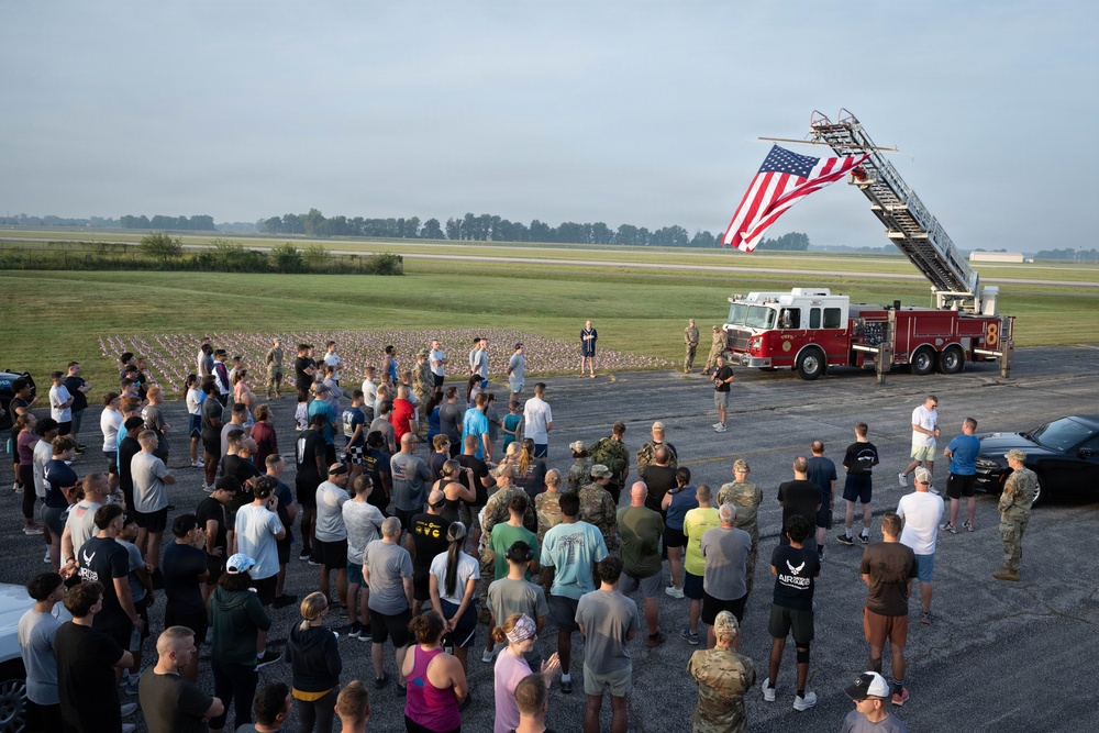 A large group of military personnel and civilians are gathered outdoors on a tarmac or airfield parking area for what appears to be a memorial or commemorative event. A red fire truck with an extended aerial ladder displays a large American flag, while a field of small flags is visible in the background. A speaker appears to be addressing the assembled crowd near the fire truck.
