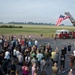A large group of military personnel and civilians are gathered outdoors on a tarmac or airfield parking area for what appears to be a memorial or commemorative event. A red fire truck with an extended aerial ladder displays a large American flag, while a field of small flags is visible in the background. A speaker appears to be addressing the assembled crowd near the fire truck.